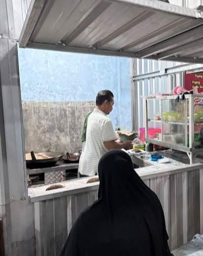 Customer in hijab watches man cooking at a simple corrugated metal food stall.