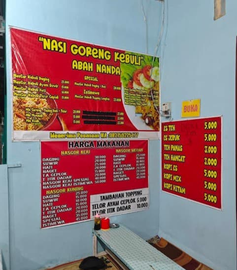 Red banners display Kebuli fried rice and drink menus in a humble Indonesian food stall.